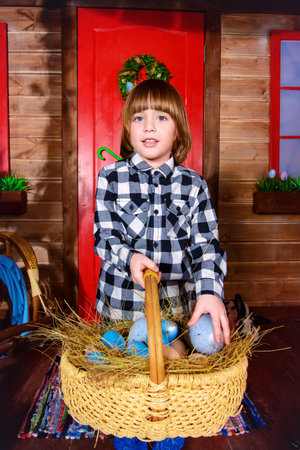 Happy little boy with basket full of painted eggs is standing on the porch near the wooden house....