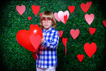 Ð¡ute child boy holding heart shaped balloon, surrounded by hearts over lawny background. First...