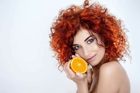Beauty, natural cosmetics and hair concept. Beautiful young woman with bright red curly hair and healthy shiny skin holding an orange. White background.の写真素材