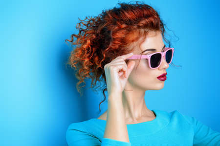 Portrait of a beautiful young woman in sunglasses and blue dress with curly foxy hair over blue background. Beauty, fashion concept. Make-up and cosmetics. Studio shot.の写真素材