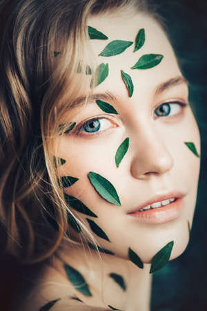Beautiful young woman with green leaves on her face over green background.の写真素材