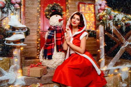 Pretty Santa girl is posing with a snowman near the house of Santa, decorated with festive lights. Christmas and New Year concept.の写真素材