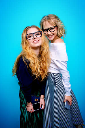 Two pretty young women dressed in sweaters and skirts posing in the studio over blue background. Winter fashion.の写真素材