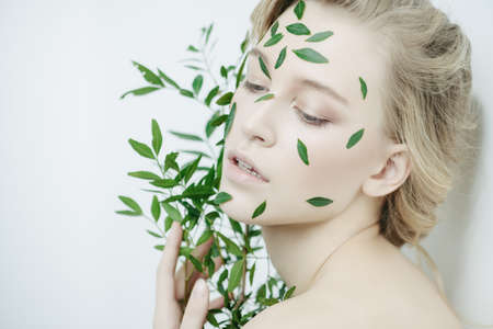 Beautiful young woman with green leaves on her face over white background. Cosmetics and makeup.の写真素材