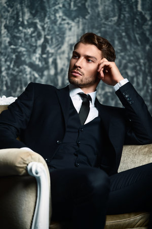 Portrait of a handsome man in an elegant suit sitting in an armchair on a grunge background. Studio...