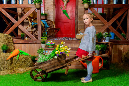 Cute little girl with a hand trolley standing in front of a wooden house. Spring. Easter holiday. の写真素材