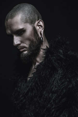 Portrait of a bearded hipster man wearing black fur coat with piercing in the ears and lips. Studio shot over dark background.の写真素材