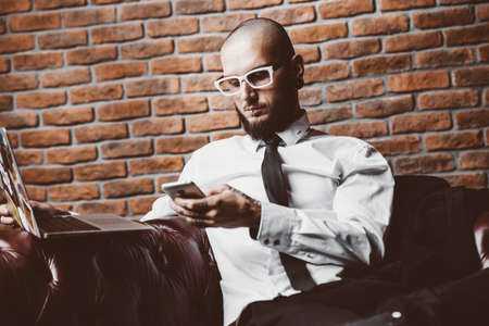 Young brutal businessman is working at home using his laptop and smartphone. Male beauty, fashion. Business concept. Modern technologies.の写真素材
