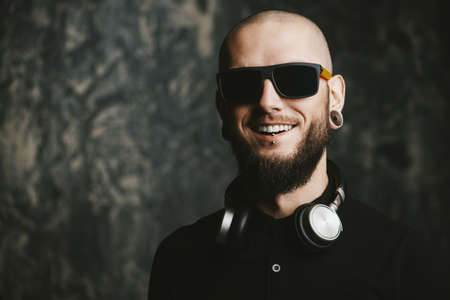 Close-up portrait of a bearded hipster man wearing sunglasses and headphones with piercing in the ears and lips. Studio shot over grunge background.の写真素材