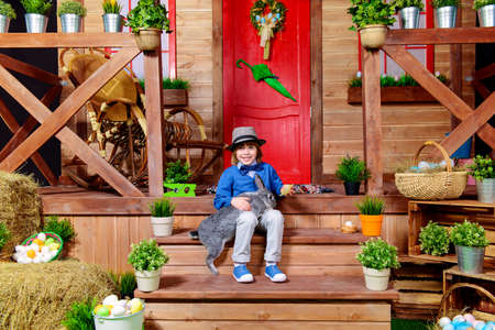 Happy little boy with bunny sitting on the porch near the wooden house. Easter holiday. Rural style, easter decoration.の写真素材