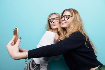 Two pretty girls are making selfie. Modern lifestyle. Studio shot over blue background.の写真素材