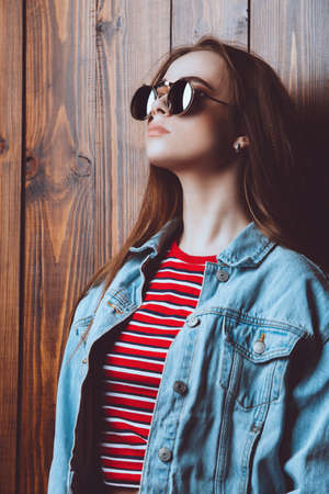 Attractive young woman in denim jacket, trendy sunglasses and red t-shirt stands by a wooden wall. の写真素材