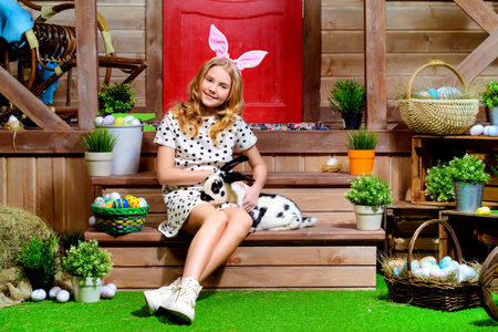 Happy little girl with bunny sitting on the porch near the wooden house. Easter holiday. Rural...