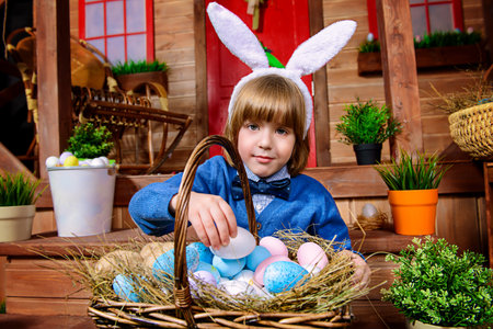 Happy little boy with basket full of painted eggs is sitting on the porch near the wooden house....