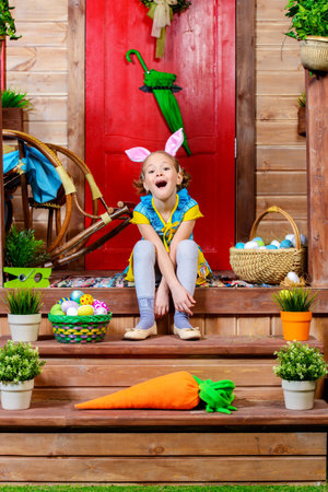 Happy little girl with bunny ears sitting on the porch of a wooden house. Easter holiday. Rural...