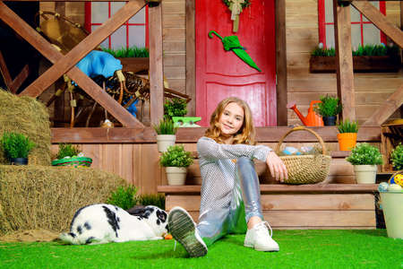 Easter holidays. Happy child girl with painted eggs is sitting on the lawn near the wooden house.の写真素材