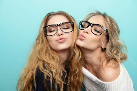 Two pretty cheerful girls in glasses are posing in studio over blue background. Beauty, fashion. Optics, eyewear.の写真素材