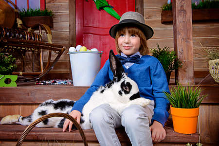 Happy little boy with bunny sitting on the porch near the wooden house. Easter holiday. Rural style, easter decoration.の写真素材