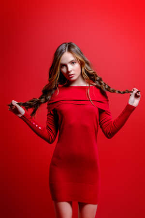 Fashion shot. Beautiful female model with long curly hair posing at studio in red dress. Red background. Beauty, fashion. の写真素材
