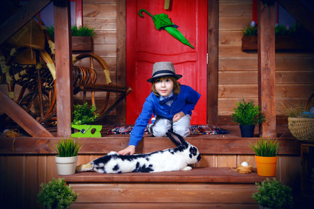 Happy little boy with bunny sitting on the porch near the wooden house. Easter holiday. Rural style,...