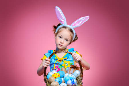 Easter holiday. Portrait of a happy little girl wearing bunny ears with basket full of painted eggs over pink background.の写真素材
