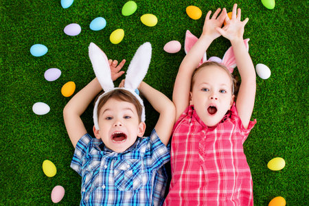 Two funny kids with bunny ears lie on a grass among colorful Easter eggs. Easter holiday.