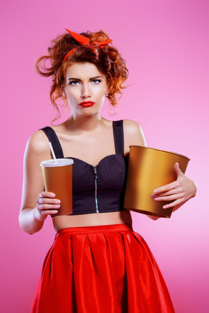 Pretty emotional young woman eating popcorn and drinking soda over pink background. Pin-up style....