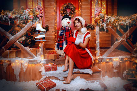 Pretty Santa girl is posing with a snowman near the house of Santa, decorated with festive lights. Christmas and New Year concept.の写真素材