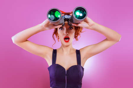Pretty red-haired girl looks through binoculars at something over pink background. Pin-up style. の写真素材