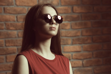 Attractive young woman in sunglasses and red t-shirt stands by a brick wall. Beauty, fashion concept.の写真素材