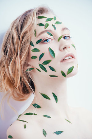 Beautiful young woman with green leaves on her face  and body over white background. Cosmetics and...