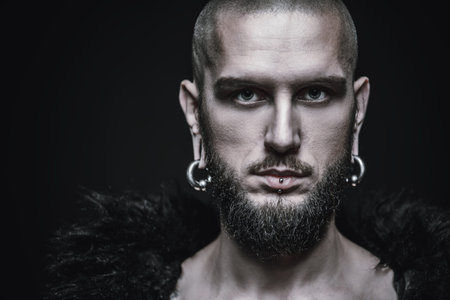 Portrait of a bearded hipster man wearing black fur coat with piercing in the ears and lips. Studio...