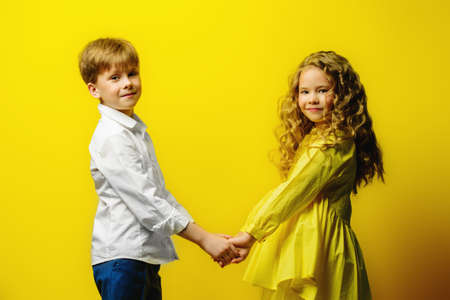 Children's fashion. Beautiful boy and girl in elegant clothes posing together at studio over yellow background.の写真素材