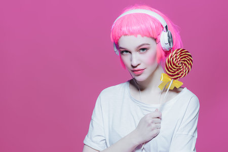 Trendy girl with pink hair wearing headphones is eating lollipop. Pink background. Youth style,...