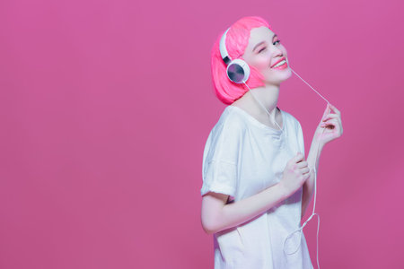 Trendy girl with pink hair enjoys the music on headphones. Pink background. Youth style, leisure.の写真素材