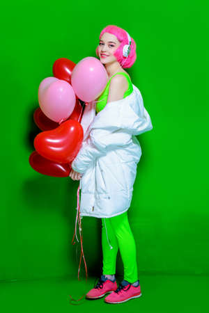 Full length portrait of a trendy girl with pink hair wearing bright stylish clothes holding balloons. Green background. Beauty, fashion, youth style.の写真素材