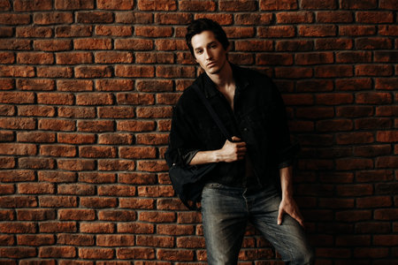 Male beauty, fashion. Handsome young man posing in black denim jacket by the brick wall. Hair...