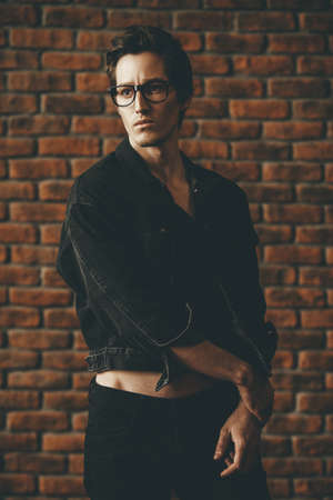 Male beauty, fashion. Handsome young man posing in black denim jacket by the brick wall. Hair styling.の写真素材