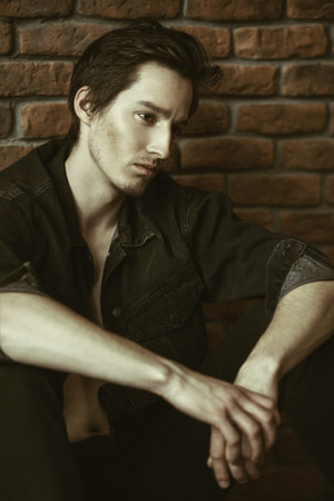 Male beauty, fashion. Handsome young man posing in black denim jacket by the brick wall. Hair...