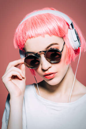 Trendy girl with pink hair wearing sunglasses enjoys the music on headphones. Pink background. Youth style, leisure.の写真素材
