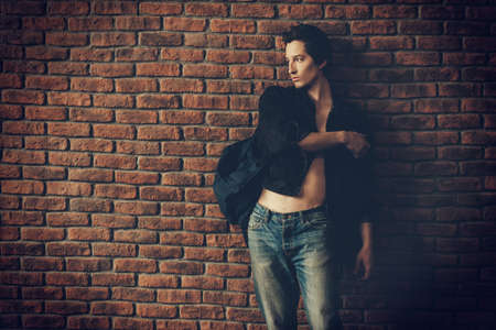 Male beauty, fashion. Handsome young man posing in black denim jacket by the brick wall. Hair styling.の写真素材