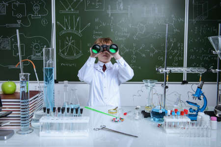 Schoolboy looks through binoculars by a chalkboard on a chemistry lesson. Educational concept.の写真素材