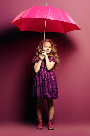 Cute little girl in a beautiful summer dress with umbrella posing at studio over purple background. Kid's fashion.の写真素材