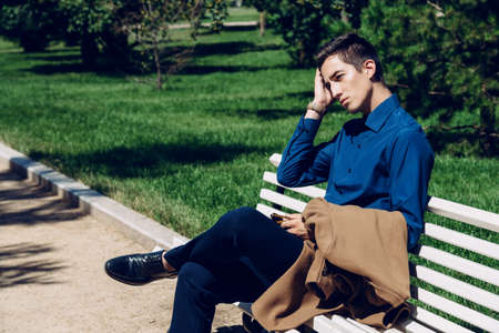 Handsome imposing young man is sitting on a bench in a park. Fashion shot. Business style. の写真素材