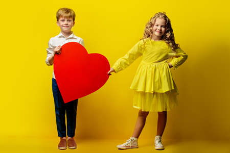 Children's fashion. Beautiful boy and girl in elegant clothes holding big red heart over yellow background. Full length portrait.の写真素材