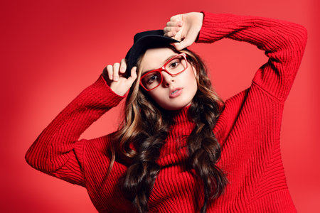 Teen style. Beautiful female model with long hair posing at studio in youth clothes. Red background....
