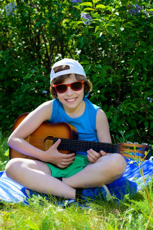 Happy ten year old boy playing guitar and singing a song in the park. Sunny summer holidays.