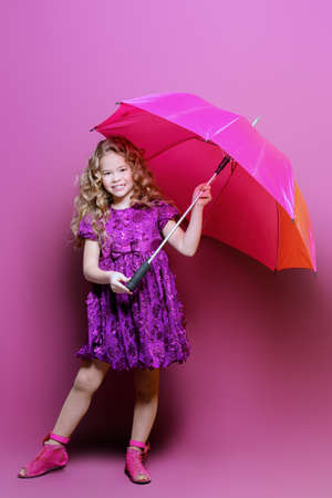 Cute little girl in a beautiful summer dress with umbrella posing at studio over purple background. Kid's fashion.の写真素材