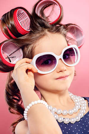 Portrait of a pretty little girl with curlers in her hair in sunglasses. Studio shot over pink background. Kid's fashion. の写真素材