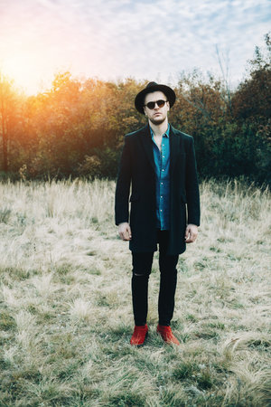 Autumn fashion. Handsome male model in black coat, a hat and sunglasses posing outdoor. Autumn...